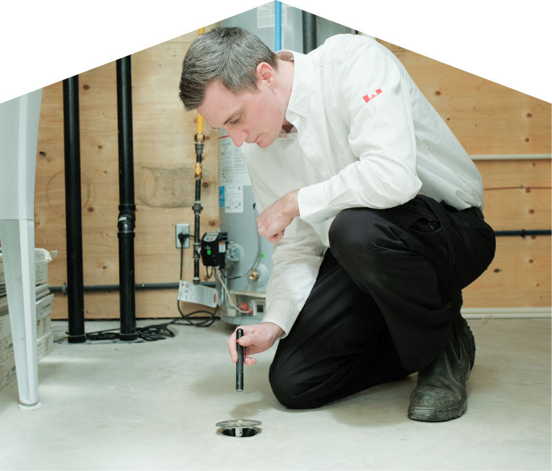 Moore & Russell plumbing specialist inspecting a basement floor drain with a flashlight during a drain cleaning service, in a utility room with visible pipes, water heater, and wood panel walls.