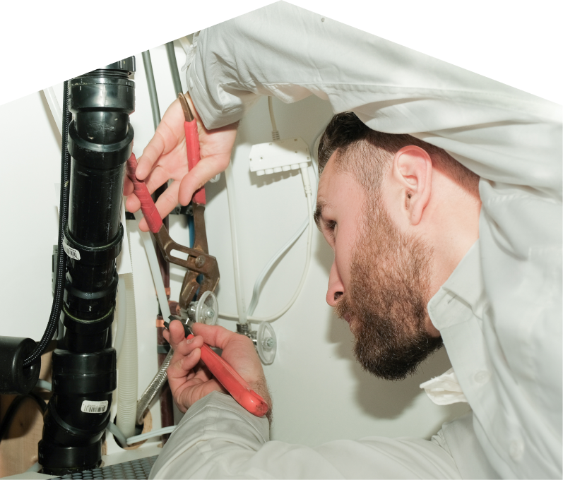 Moore & Russell plumbing specialist uses hand tools to tighten pipe fittings beneath a sink during a repair, with visible wiring and drainage lines in the background. Moore & Russell plumbing specialist uses hand tools to tighten pipe fittings beneath a sink during a repair, with visible wiring and drainage lines in the background.