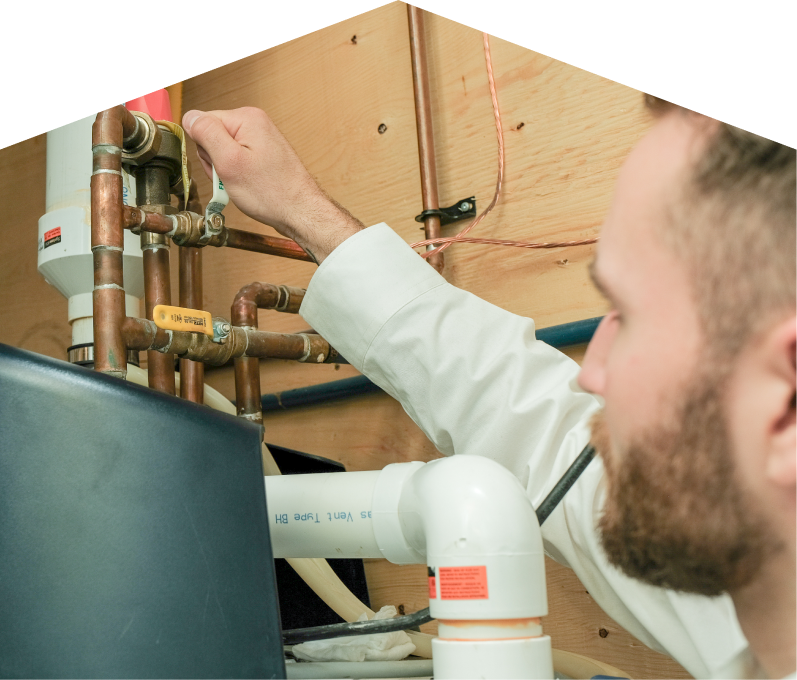 Moore & Russell water heater specialist tightens a valve on copper piping during a water heater installation, with exposed wood paneling and wiring in the background. Moore & Russell water heater specialist tightens a valve on copper piping during a water heater installation, with exposed wood paneling and wiring in the background.